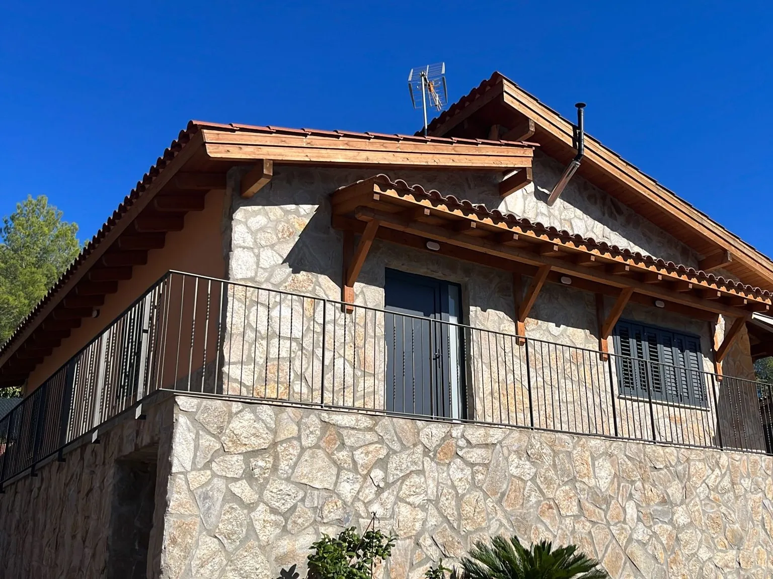 Casa amb la façana i el mur revestits de pedra, teulada de teula amb ràfecs de fusta i barana negra